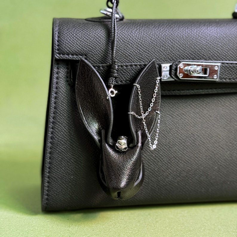 Goth luxury bunny leather bag charm hanging on a black designer handbag, handmade lambskin wicked purse charm with long ears and metal chain accents. Dark gothic handbag charm designed as a statement accessory, black leather bunny bag charm for handbags, purses, and luxury bags, showcasing minimalist dark luxury style and edgy aesthetic.