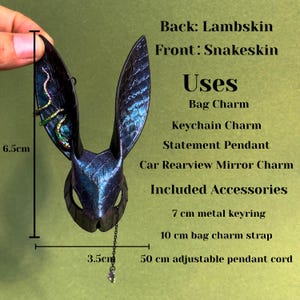 A handmade bunny-shaped leather bag charm hanging on a textured black handbag. The charm is crafted from blue-purple rainbow snakeskin on the front with lambskin backing, featuring tall pointed ears, embossed reptile scales, and an iridescent reflective surface. A small decorative chain with a crystal drop hangs from the chin, and the ear includes a colorful snake-shaped accent. The background is light green.