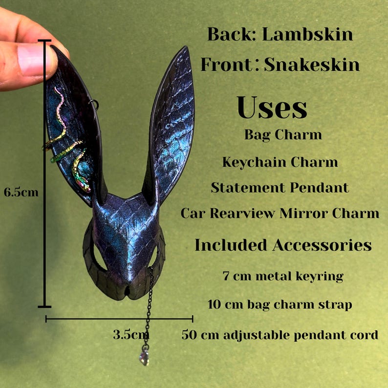 A handmade bunny-shaped leather bag charm hanging on a textured black handbag. The charm is crafted from blue-purple rainbow snakeskin on the front with lambskin backing, featuring tall pointed ears, embossed reptile scales, and an iridescent reflective surface. A small decorative chain with a crystal drop hangs from the chin, and the ear includes a colorful snake-shaped accent. The background is light green.