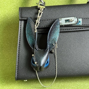 May include: A black leather handbag with a silver chain and clasp. A decorative bat-shaped charm with iridescent teal and blue wings and a blue accent is attached to the bag with a silver chain.