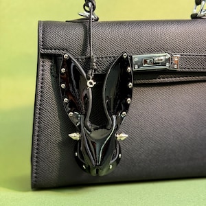 Puede incluir: Un dije de conejito negro brillante con tachuelas y púas plateadas cuelga de un bolso de mano de cuero negro. El dije de conejito tiene orejas largas y una cara detallada. El bolso es negro con herrajes plateados.
