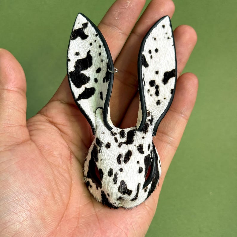 Pode incluir: Broche em forma de orelhas de coelho com manchas pretas sobre fundo branco. O broche &eacute; feito de um material texturizado. Os contornos s&atilde;o pretos. Um acess&oacute;rio &uacute;nico para roupas.