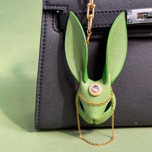 Puede incluir: Un charm de bolso con forma de conejo de cuero verde, con largas orejas y una cadena dorada. El charm tiene una joya decorativa en el centro y está sujeto a un bolso de mano de cuero negro. El fondo es verde claro.