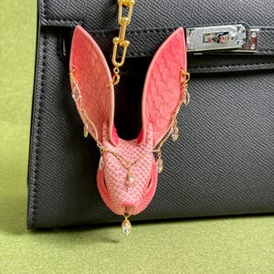 May include: A pink and red leather bunny charm with gold chain and crystal accents. The bunny charm is attached to a black leather handbag. The bunny has long ears and is decorated with gold chains and dangling crystals.