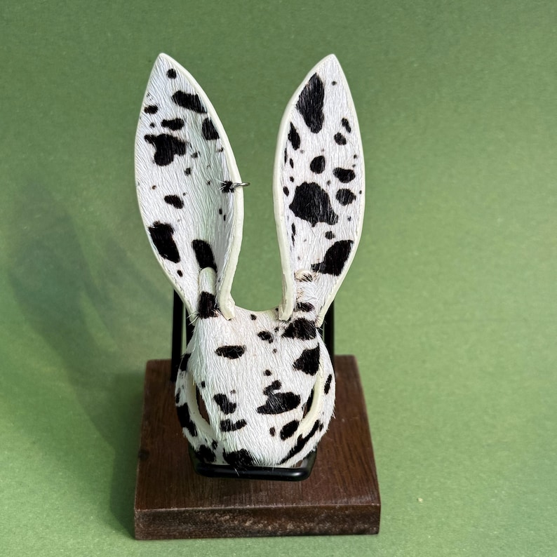 Pode incluir: Uma escultura decorativa de coelho com um padr&atilde;o de manchas em preto e branco. As orelhas do coelho est&atilde;o em p&eacute; e a escultura &eacute; montada em uma pequena base de madeira escura. O fundo &eacute; verde liso.