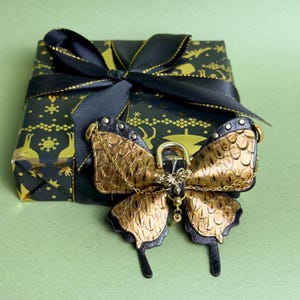 May include: A decorative butterfly-shaped object with gold and black accents, featuring a textured pattern. The butterfly is adorned with a gold chain and is placed on top of a wrapped gift box with a black bow. The gift box is wrapped in black paper with gold designs.