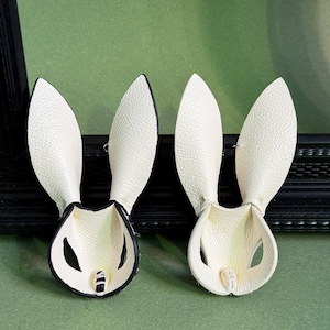 Pode incluir: Dois acess&oacute;rios de cabelo em forma de orelhas de coelho em couro branco. As orelhas t&ecirc;m acabamento preto e est&atilde;o sobre um fundo verde. Estes acess&oacute;rios s&atilde;o projetados para serem usados na cabe&ccedil;a.