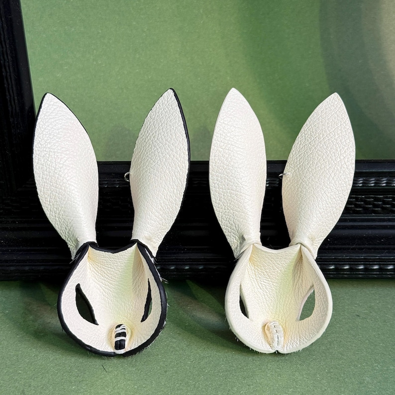 Pode incluir: Dois acess&oacute;rios de cabelo em forma de orelhas de coelho em couro branco. As orelhas t&ecirc;m acabamento preto e est&atilde;o sobre um fundo verde. Estes acess&oacute;rios s&atilde;o projetados para serem usados na cabe&ccedil;a.