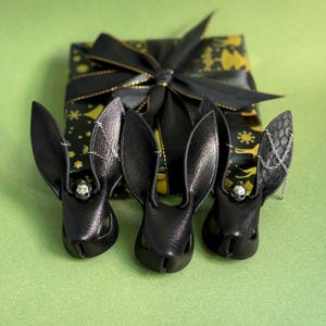 Op de afbeelding: Drie zwarte leren konijnenoorhangers met schedelaccenten en zilveren kettingen, geplaatst voor een cadeauverpakking. De cadeauverpakking is zwart met een gouden patroon en een zwarte strik.