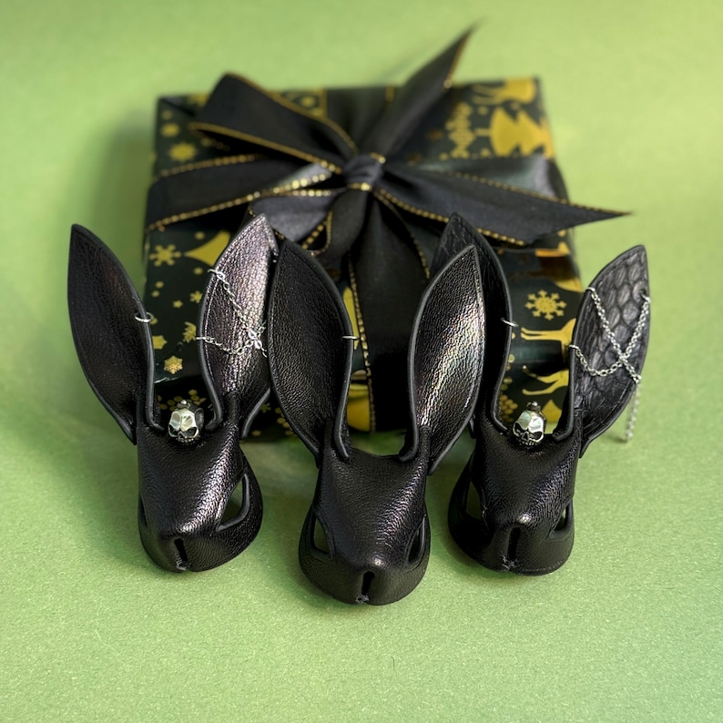 Op de afbeelding: Drie zwarte leren konijnenoorhangers met schedelaccenten en zilveren kettingen, geplaatst voor een cadeauverpakking. De cadeauverpakking is zwart met een gouden patroon en een zwarte strik.
