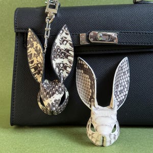 May include: Two rabbit-shaped keychains made from reptile skin, featuring a pattern of black and cream scales. One keychain is attached to a black leather handbag with silver hardware. The rabbit keychains have long, upright ears and detailed facial features.