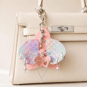 May include: A decorative keychain charm with a pink and iridescent wing design and silver details. The charm is attached to a white handbag. The wings have a scale-like pattern with shades of pink, blue, and purple.