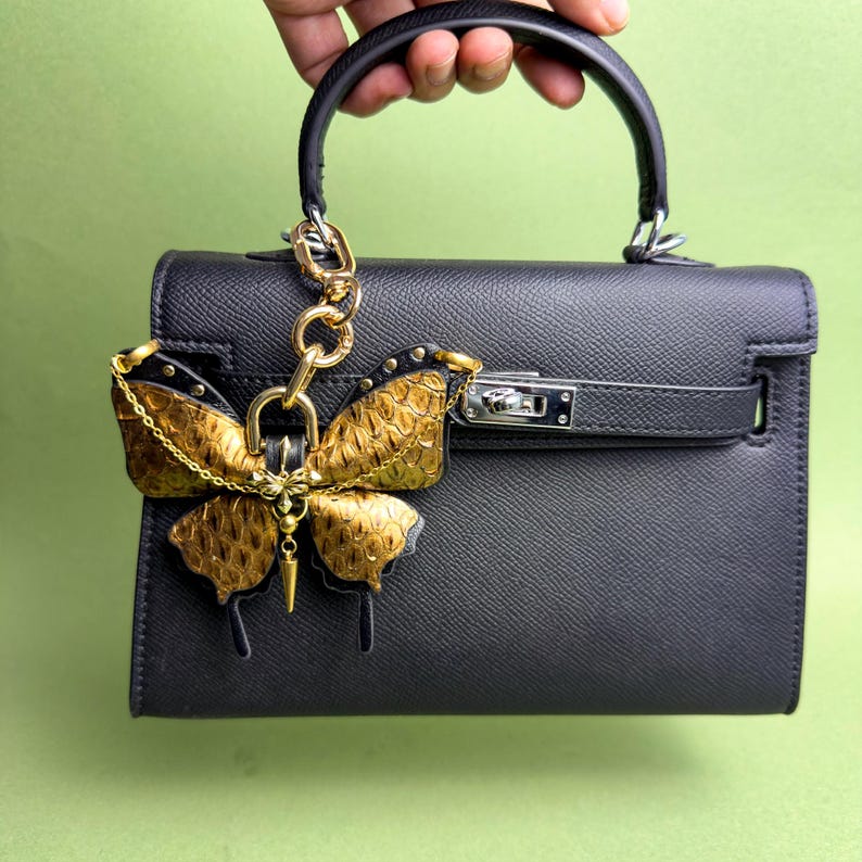 Gold vintage gothic butterfly leather bag charm handmade from luxury snakeskin and lambskin leather, shown attached to a structured black handbag held by hand to demonstrate size and proportion. This leather butterfly purse charm features gold-tone metal hardware, chain accents, and a rich gold snakeskin texture, designed as a statement handbag charm with a dark luxury vintage gothic aesthetic.