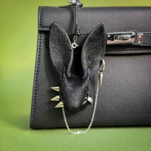 May include: A black leather handbag with a decorative black rabbit head charm. The rabbit charm has silver spikes and a silver chain. The handbag has a silver clasp and a black leather strap.