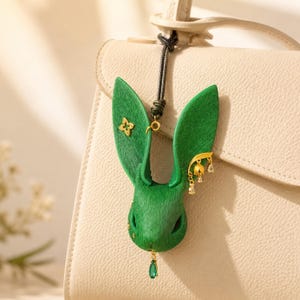 Puede incluir: Un charm de bolso con forma de conejo verde con detalles dorados. El charm presenta un adorno en forma de estrella en una oreja y colgantes dorados y de cristal. Una gema verde cuelga de la parte inferior. El charm está sujeto a un cordón negro.
