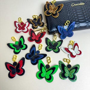 A collection of handmade leather wallet charms in multiple colors, all shaped as layered butterflies crafted from genuine snakeskin and French lambskin. The charms are shown together around a black textured wallet to display their size and color options. Each leather purse charm features gold-plated hardware and can be used as a handbag charm or zipper charm. This image highlights the available style choices for shoppers.