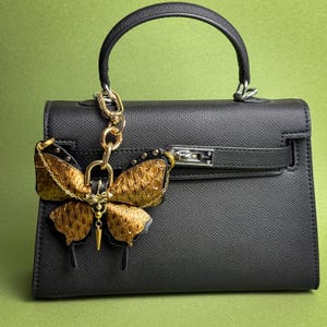 Gold vintage gothic butterfly leather bag charm handmade from luxury snakeskin and lambskin leather, displayed attached to a structured black handbag to show overall size and styling. This leather butterfly purse charm features gold-tone metal hardware, chain details, and a rich gold snakeskin texture, designed as a statement handbag charm with a dark luxury vintage gothic style.