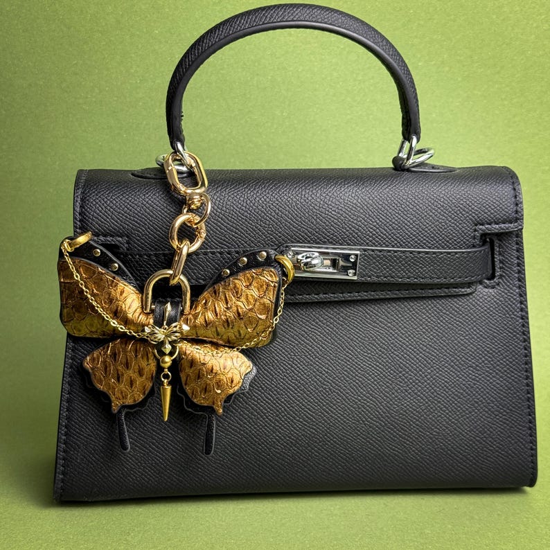 Gold vintage gothic butterfly leather bag charm handmade from luxury snakeskin and lambskin leather, displayed attached to a structured black handbag to show overall size and styling. This leather butterfly purse charm features gold-tone metal hardware, chain details, and a rich gold snakeskin texture, designed as a statement handbag charm with a dark luxury vintage gothic style.