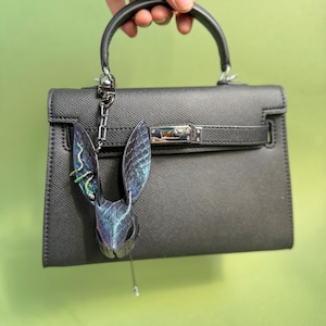 Blue-purple rainbow snakeskin bunny leather bag charm hanging from a black leather handbag; the leather purse charm features iridescent snakeskin texture with shifting blue, purple, and rainbow tones, lambskin backing, long sculpted ears, rainbow accent snake detail on one ear, and a dangling teardrop pendant. The handmade leather bag charm is shown attached to the handbag metal hardware, highlighting shine and color shifts.