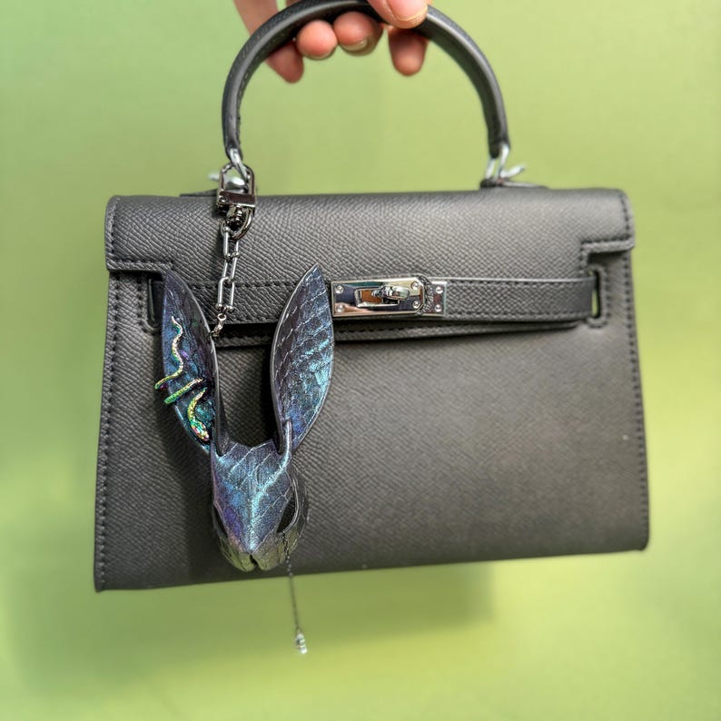 Blue-purple rainbow snakeskin bunny leather bag charm hanging from a black leather handbag; the leather purse charm features iridescent snakeskin texture with shifting blue, purple, and rainbow tones, lambskin backing, long sculpted ears, rainbow accent snake detail on one ear, and a dangling teardrop pendant. The handmade leather bag charm is shown attached to the handbag metal hardware, highlighting shine and color shifts.