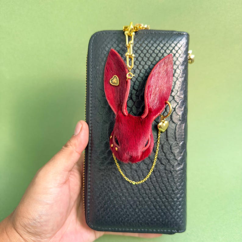 Burgundy Gothic bunny leather bag charm displayed on a black snakeskin wallet, featuring handmade horsehair leather, lambskin backing, gold heart accents, and a delicate chain. Luxury Dark Romance purse charm suitable for handbags, wallets, and designer accessories.