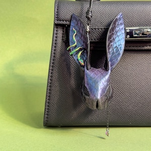 Blue-purple rainbow snakeskin bunny leather bag charm hanging on a black handbag; handmade lambskin & snakeskin leather purse charm with iridescent blue-purple finish, long sculpted ears, rainbow accents on one ear, and a small dangling crystal; suitable for leather bag charm, leather purse charm, keychain charm, or handbag charms display.