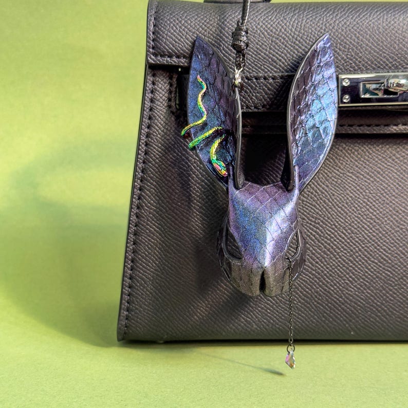 Blue-purple rainbow snakeskin bunny leather bag charm hanging on a black handbag; handmade lambskin & snakeskin leather purse charm with iridescent blue-purple finish, long sculpted ears, rainbow accents on one ear, and a small dangling crystal; suitable for leather bag charm, leather purse charm, keychain charm, or handbag charms display.