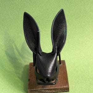 Op de afbeelding: Een zwart leren konijnenmasker met lange, rechtopstaande oren. Het masker heeft uitsparingen voor ogen en neus en is gemonteerd op een houten voet met een metalen standaard. Het masker wordt gepresenteerd tegen een groene achtergrond.