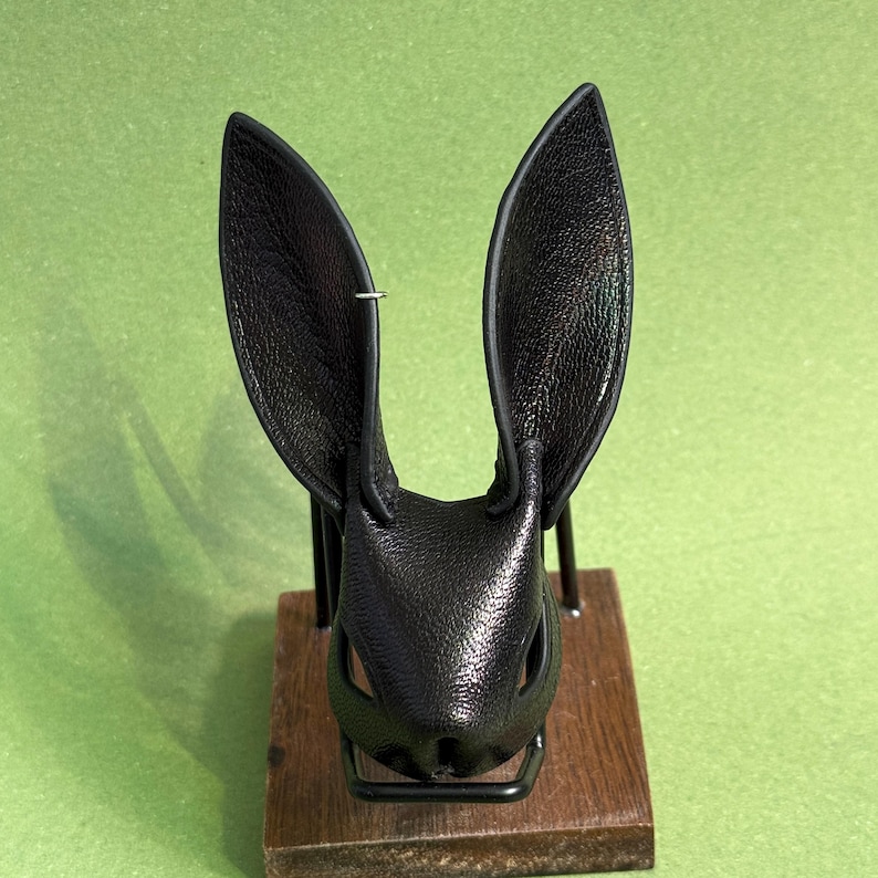 Op de afbeelding: Een zwart leren konijnenmasker met lange, rechtopstaande oren. Het masker heeft uitsparingen voor ogen en neus en is gemonteerd op een houten voet met een metalen standaard. Het masker wordt gepresenteerd tegen een groene achtergrond.