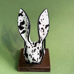 Pode incluir: Escultura decorativa de cabe&ccedil;a de coelho com orelhas longas. O padr&atilde;o apresenta manchas pretas sobre fundo branco, semelhante a um d&aacute;lmata. As orelhas s&atilde;o contornadas em preto e eretas. A escultura est&aacute; montada numa pequena base de madeira escura.