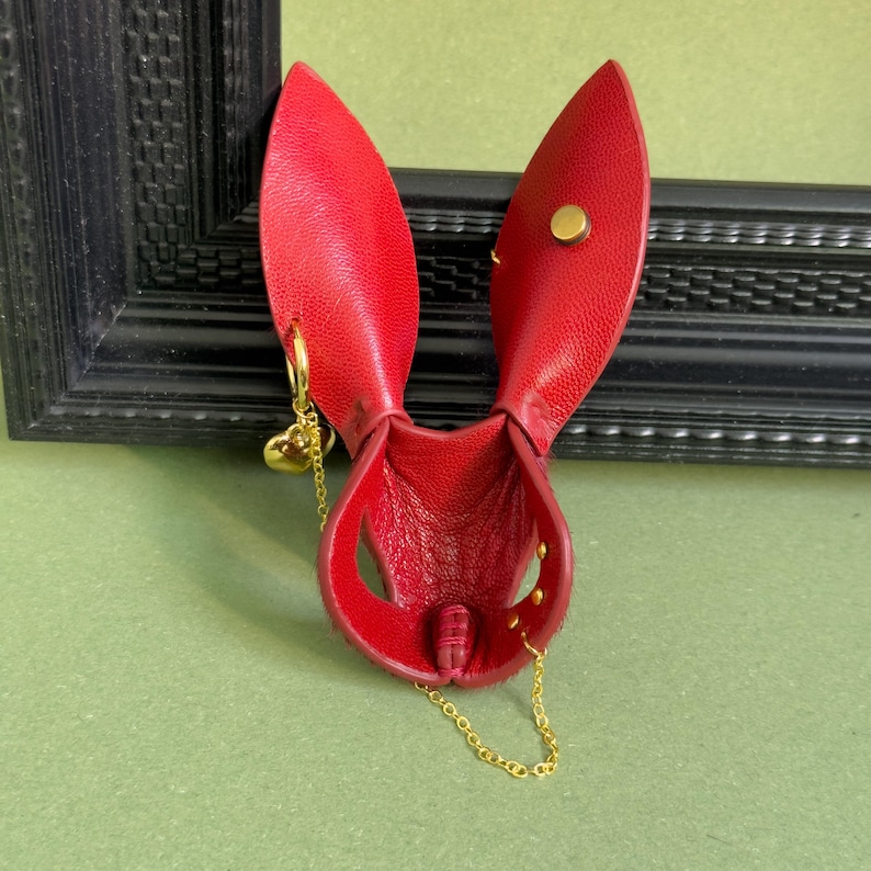 May include: A red leather pouch with a bunny ear design. The pouch has a gold chain and a small gold heart charm. The pouch is secured with a gold snap and rivets.
