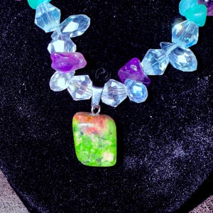 May include: A colorful necklace featuring a pendant with green and red hues, complemented by clear, purple, and turquoise beads. The necklace is displayed against a black background, highlighting the vibrant colors of the gemstones.