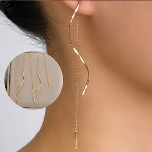 Puede incluir: Pendientes largos dorados. Un pendiente se lleva puesto, mostrando un diseño fluido y curvado. Los otros dos pendientes se muestran en un estuche circular. Los pendientes tienen una delicada cadena y una barra recta en el extremo.