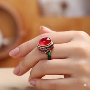 Puede incluir: Primer plano de un anillo con una gema roja grande y redonda. El anillo tiene una banda dorada con detalles decorativos azules y rojos. Un pequeño detalle verde es visible en el vástago del anillo.