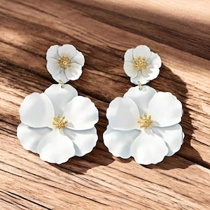 Puede incluir: Un par de pendientes blancos con forma de flor y centros dorados. Los pendientes tienen un diseño de pétalos en capas y están colocados sobre un fondo de madera. Son una pieza llamativa.