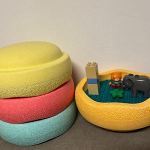 Può includere: Una pila di tre oggetti rotondi, strutturati e colorati in giallo, corallo e turchese. Accanto, una ciotola gialla con un tappetino blu, un orsetto giocattolo, un elefante grigio e un mattoncino Lego.