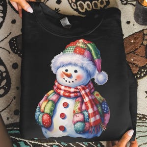 May include: Black sweatshirt featuring a watercolor illustration of a snowman. The snowman has a patchwork scarf, hat, and body. The snowman has a carrot nose, button eyes, and a red button mouth. The snowman is wearing a red and white plaid scarf.