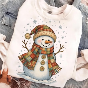 May include: White sweatshirt featuring a snowman illustration. The snowman has a carrot nose, twig arms, and a plaid scarf and hat. Three decorative buttons are on the snowman's body. Snowflakes are scattered around the snowman.