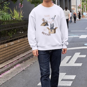 Op de afbeelding: Een witte sweatshirt met een afbeelding met de tekst "Things Can Only Get Better". De afbeelding toont een kat in een doos, een duif en een nijlpaard. De persoon draagt een donkerblauwe jeans en zwart-witte sneakers.