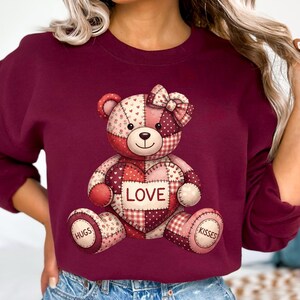 May include: Burgundy sweatshirt featuring a patchwork teddy bear design. The bear has a heart with the word "LOVE" and the words "HUGS" and "KISSES". The bear is made of red, pink, and white fabric. A bow is on the bear's head.