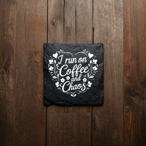 Peut inclure: Un dessous de verre carr&eacute; en ardoise noire avec un motif imprim&eacute; blanc en forme de c&oelig;ur compos&eacute; de vignes et de feuilles florales. Le texte au centre indique I run on Coffee and Chaos dans une combinaison de polices script et sans-serif.