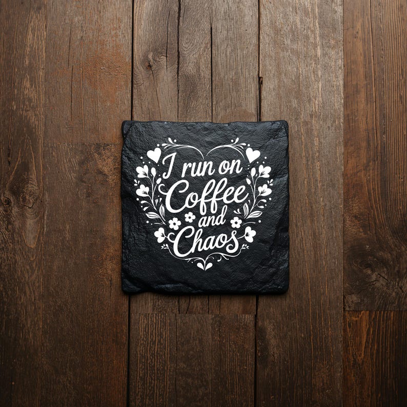 Peut inclure: Un dessous de verre carr&eacute; en ardoise noire avec un motif imprim&eacute; blanc en forme de c&oelig;ur compos&eacute; de vignes et de feuilles florales. Le texte au centre indique I run on Coffee and Chaos dans une combinaison de polices script et sans-serif.