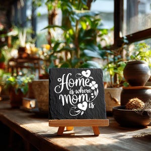 Peut inclure: Une plaque carr&eacute;e en ardoise noire avec le texte imprim&eacute; en blanc Home is where mom is, orn&eacute;e de motifs floraux et de c&oelig;urs. La plaque est pr&eacute;sent&eacute;e sur un petit chevalet en bois, mettant en valeur un bord en pierre naturelle textur&eacute;e.