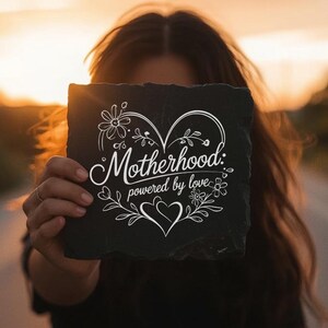 Peut inclure: Une plaque carr&eacute;e en ardoise noire avec une gravure blanche portant l'inscription Motherhood: powered by love. Le motif comprend un grand contour de c&oelig;ur, des accents floraux et de petits c&oelig;urs, le tout dans un style lin&eacute;aire blanc.