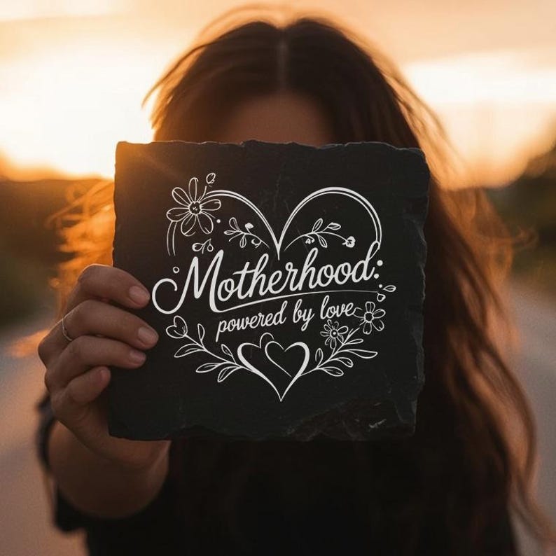 Peut inclure: Une plaque carr&eacute;e en ardoise noire avec une gravure blanche portant l'inscription Motherhood: powered by love. Le motif comprend un grand contour de c&oelig;ur, des accents floraux et de petits c&oelig;urs, le tout dans un style lin&eacute;aire blanc.