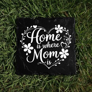 Peut inclure: Une plaque carr&eacute;e en ardoise noire avec le texte imprim&eacute; en blanc Home is where Mom is. Le texte est entour&eacute; d'un cadre d&eacute;coratif blanc en forme de c&oelig;ur compos&eacute; d'&eacute;l&eacute;ments floraux, de vignes et de petits c&oelig;urs.