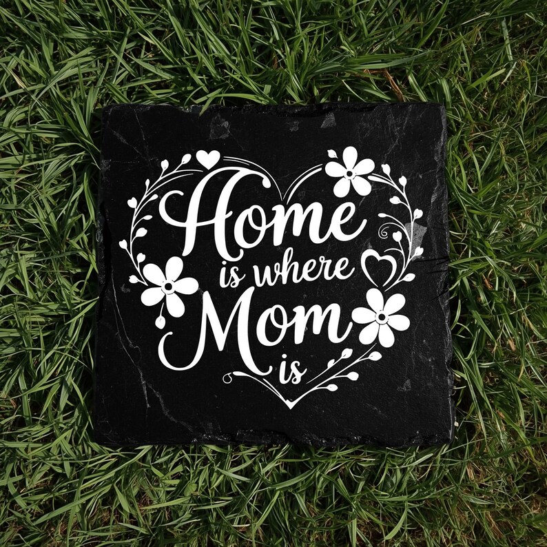 Peut inclure: Une plaque carr&eacute;e en ardoise noire avec le texte imprim&eacute; en blanc Home is where Mom is. Le texte est entour&eacute; d'un cadre d&eacute;coratif blanc en forme de c&oelig;ur compos&eacute; d'&eacute;l&eacute;ments floraux, de vignes et de petits c&oelig;urs.