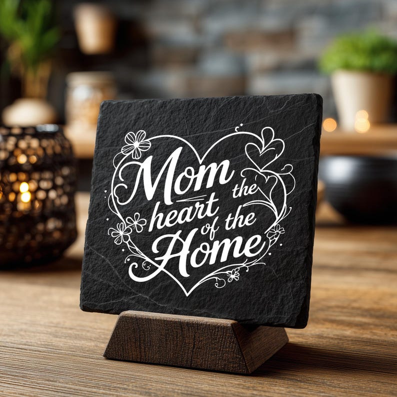 Peut inclure: Une plaque carr&eacute;e en ardoise noire &agrave; la surface rugueuse et textur&eacute;e, reposant sur un support en bois fonc&eacute;. La plaque pr&eacute;sente une inscription blanche grav&eacute;e "Mom the heart of the Home" entour&eacute;e d'un contour floral en forme de c&oelig;ur.