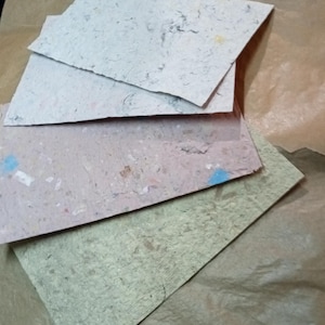 Peut inclure: Une collection de feuilles de papier faites à la main de différentes couleurs, dont vert clair, rose et blanc cassé. Le papier a une surface texturée avec des flocons et des fibres visibles, suggérant une composition naturelle et recyclée. Les feuilles sont disposées sur une surface en papier brun.