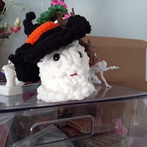 Puede incluir: Un muñeco de nieve de peluche blanco hecho a mano con un sombrero de bruja negro con una banda naranja. El muñeco de nieve tiene ojos negros de botón y una pequeña boca roja. El sombrero está adornado con ramas marrones y follaje verde.